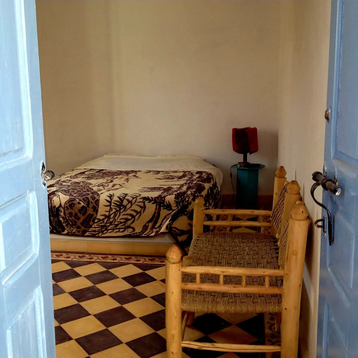 Une chambre vue depuis sa porte bleue, un lit, deux chaises en bois et corde, un sol en damier.
