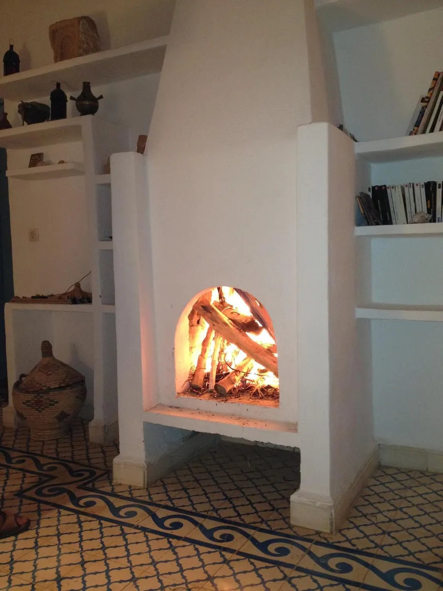 Une cheminée blanche où brûle un feu de bois, entre des étagères maçonnées garnies de livres et de poteries.
