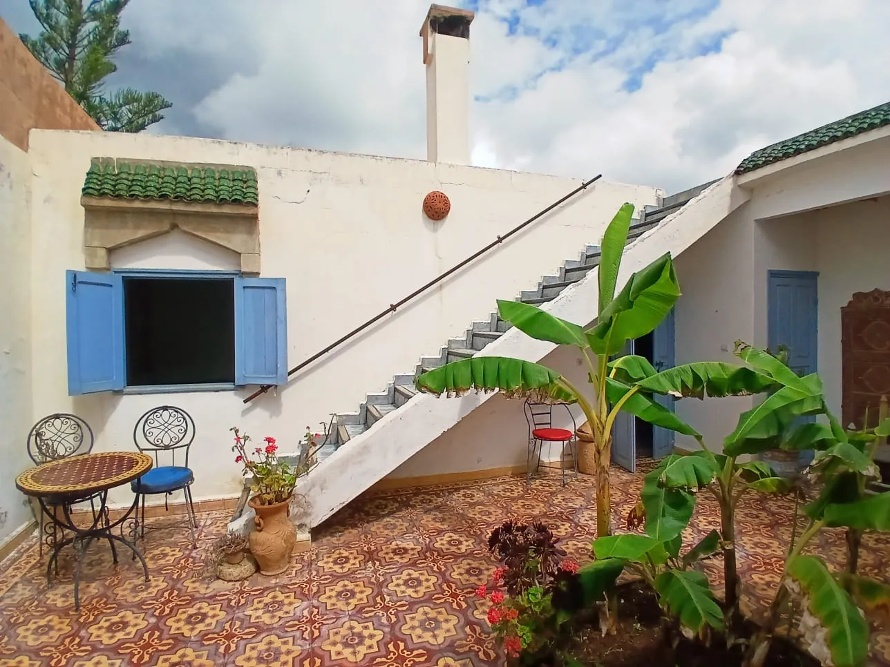 La cour carrelée de motifs, avec ses bananiers, un escalier blanc qui monte vers le toit et une fenêtre aux volets bleus.